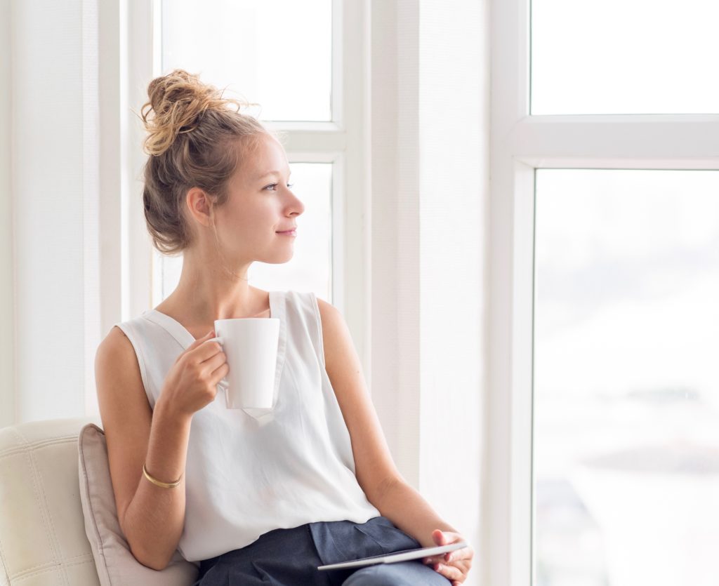 Close-up portrait of dreamy young beautiful fair-haired woman drinking tea, using tablet and looking through window on loggia at home